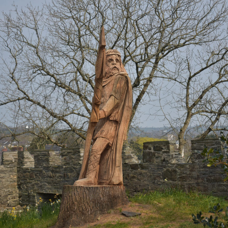 Wooden sculpture of a historical figure at Cardigan Castle, surrounded by spring blossoms and ancient walls